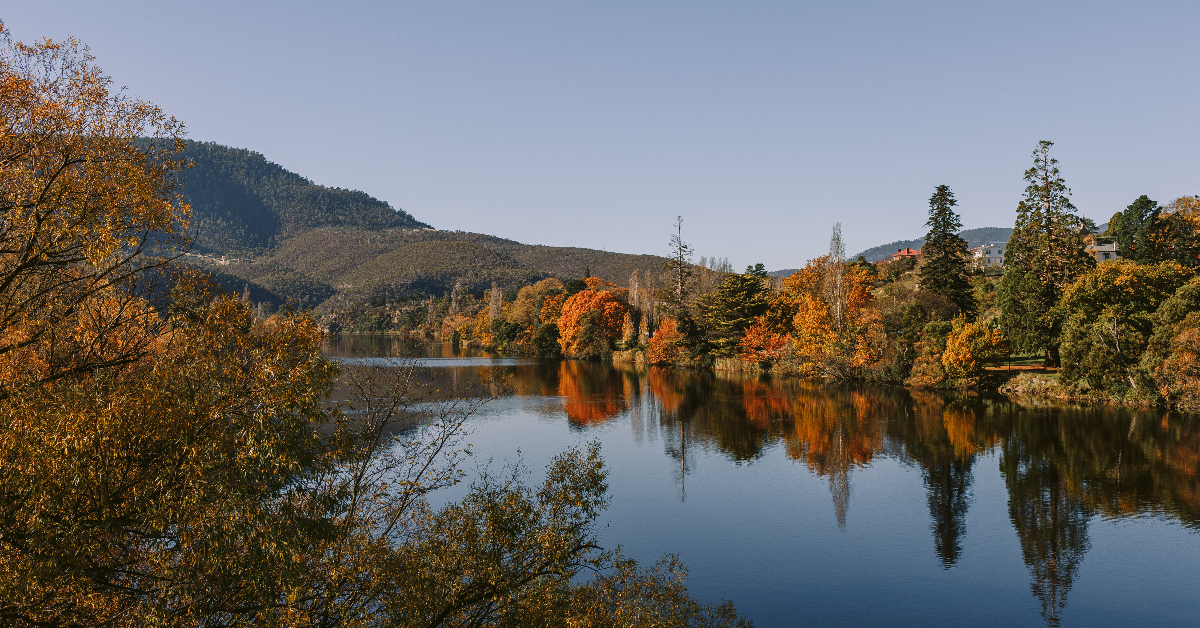 Autumn trees reflected in a calm river with hills in the background.